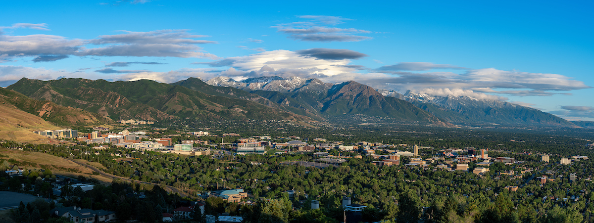 Decorative landscape photo of the university of utah campus with green trees, mountains in the background and blue sky with clouds.